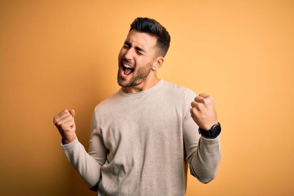 Young handsome man wearing casual sweater standing over isolated yellow background very happy and excited doing winner gesture with arms raised, smiling and screaming for success. Celebration concept.